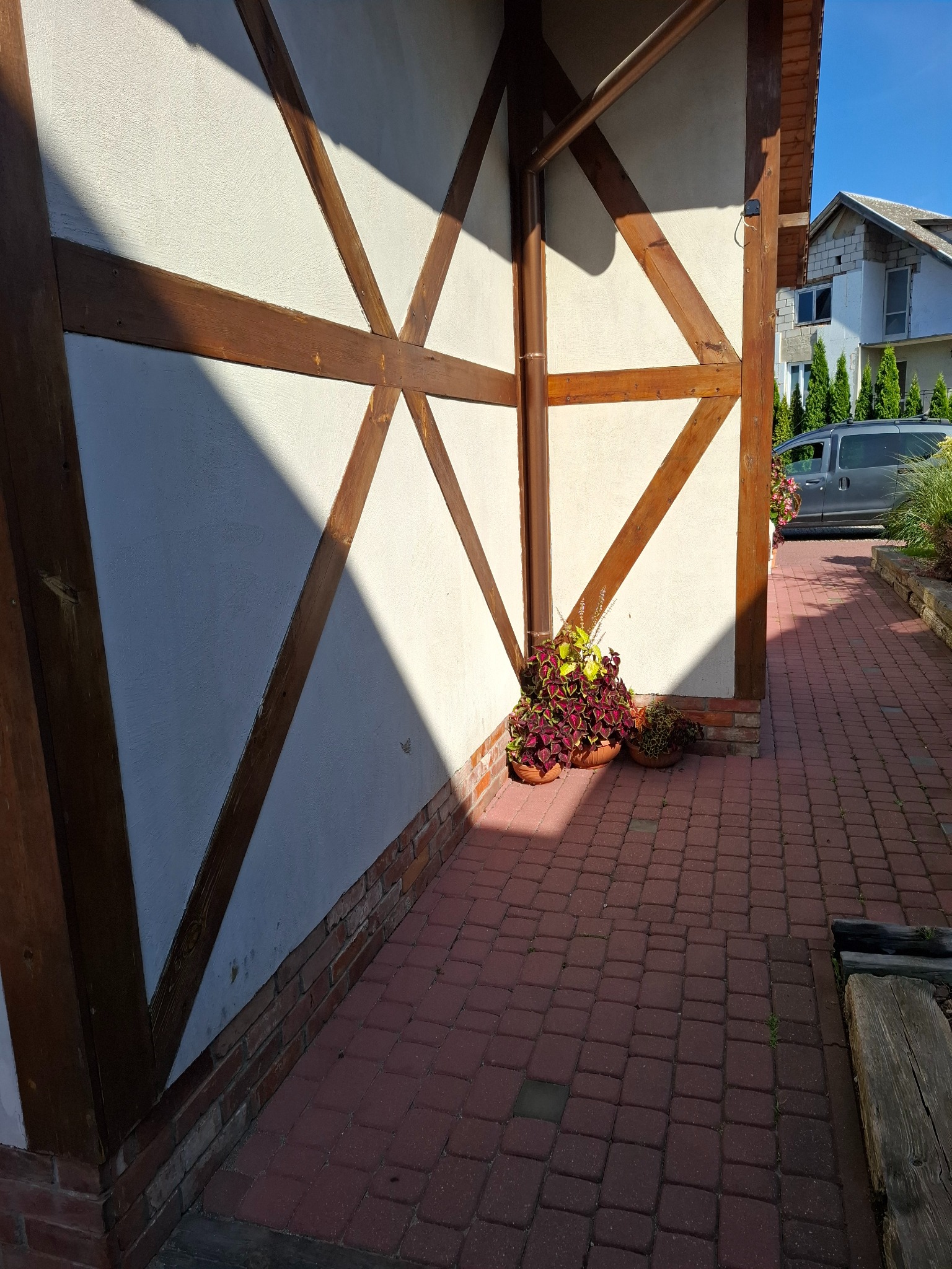 Fragment ściany budynku w stylu szachulcowym z drewnianą konstrukcją i doniczkami z kwiatami, oświetlony promieniami słońca. Czerwona kostka brukowa na ścieżce.