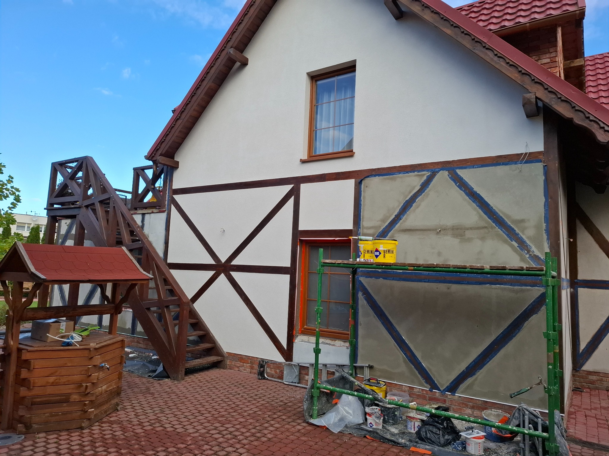 Elewacja domu w trakcie renowacji: fragment ściany z widocznym rusztowaniem, wiaderkami farby i narzędziami. Widoczna studnia z drewnianą konstrukcją.