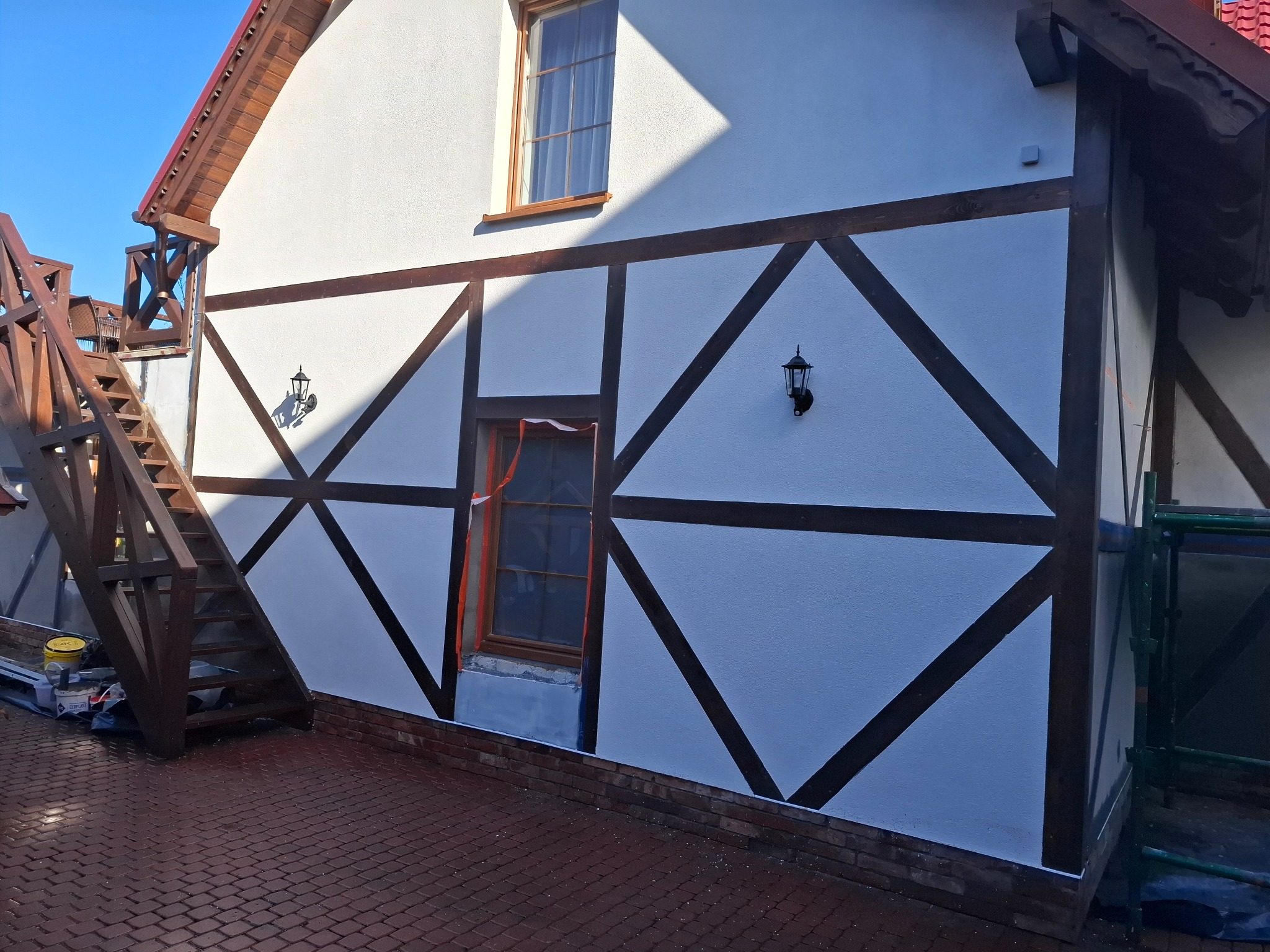 Biała elewacja domu z drewnianą konstrukcją w stylu szachulcowym, widoczne lampy zewnętrzne i fragment drewnianych schodów. Część okna zabezpieczona folią.