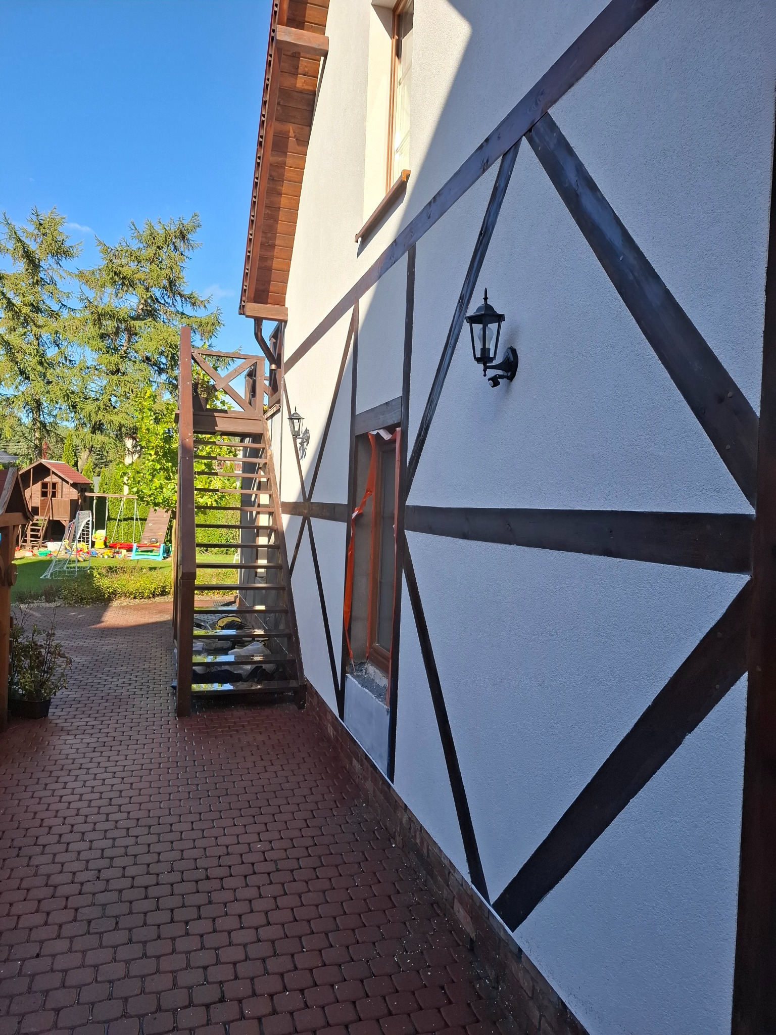 Elewacja domu z drewnianą konstrukcją szkieletową, oświetlona lampą. Widoczne schody zewnętrzne i fragment podwórka z placem zabaw w tle.