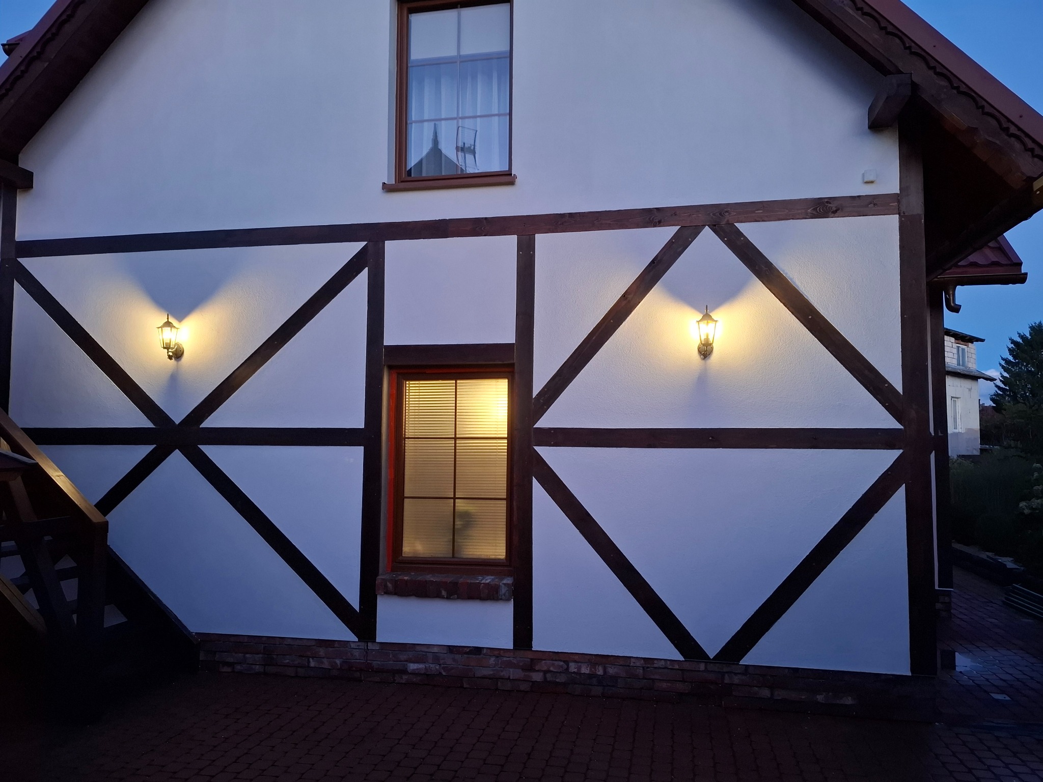 Elewacja domu w stylu szachulcowym z oknami i oświetleniem zewnętrznym. Widoczne elementy drewniane i fragment chodnika z kostki.