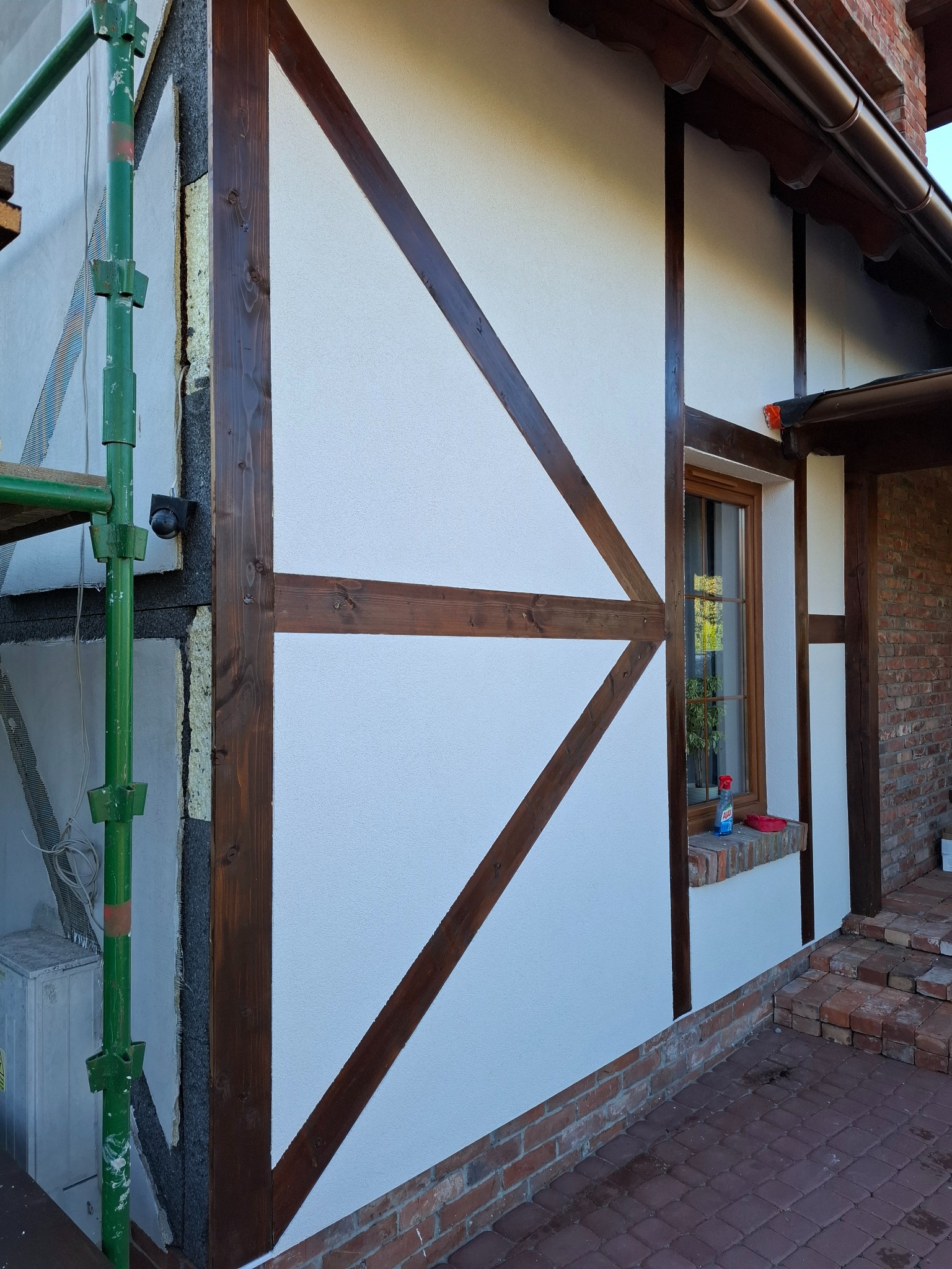 Fragment elewacji domu w stylu szachulcowym po ociepleniu i nałożeniu białego tynku. Widoczne drewniane elementy konstrukcyjne i rusztowanie z boku.