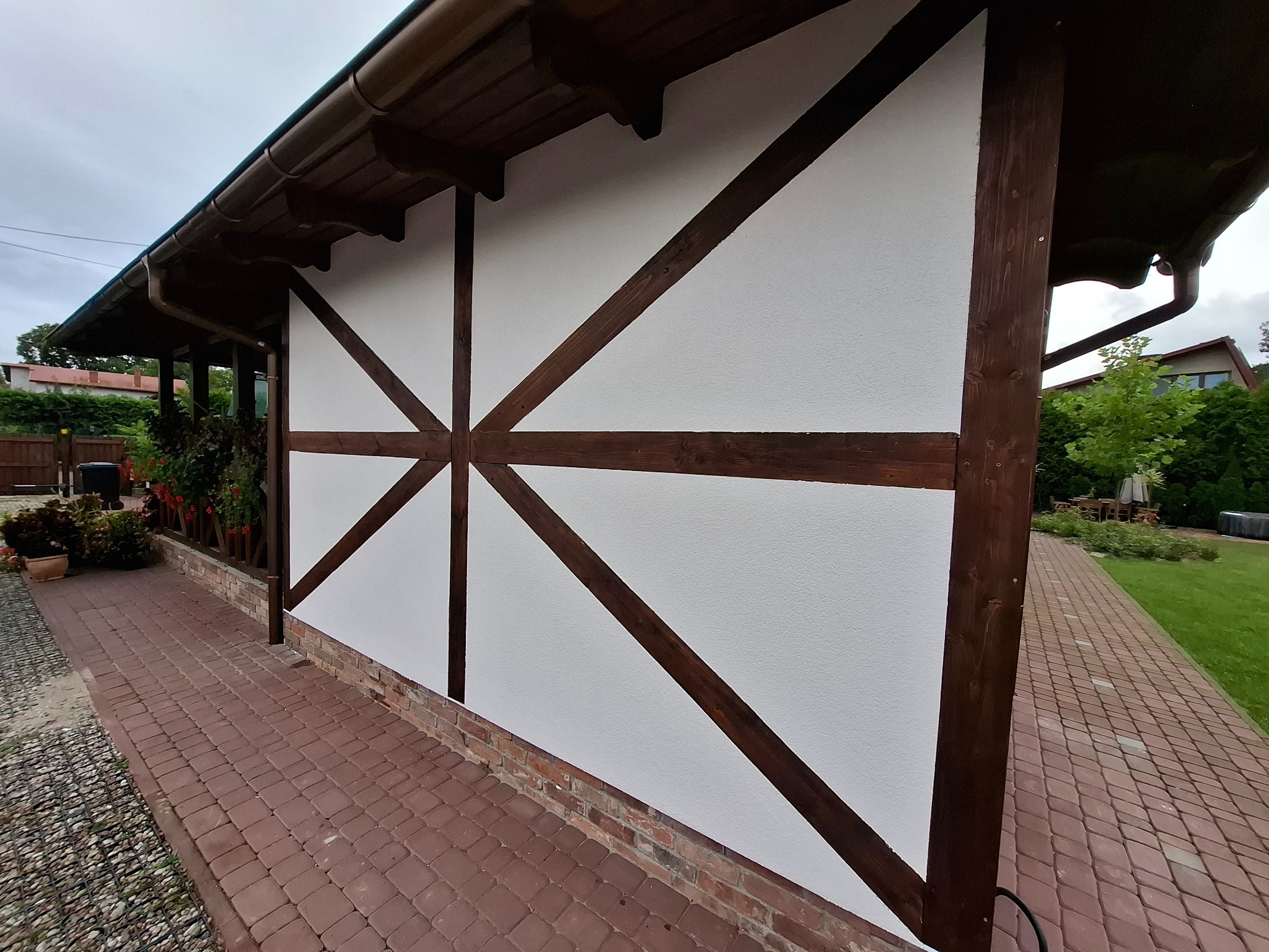 Biała elewacja budynku z drewnianą konstrukcją szkieletową w brązowym kolorze, widok z boku, z fragmentem chodnika z kostki brukowej i zieleni w tle.