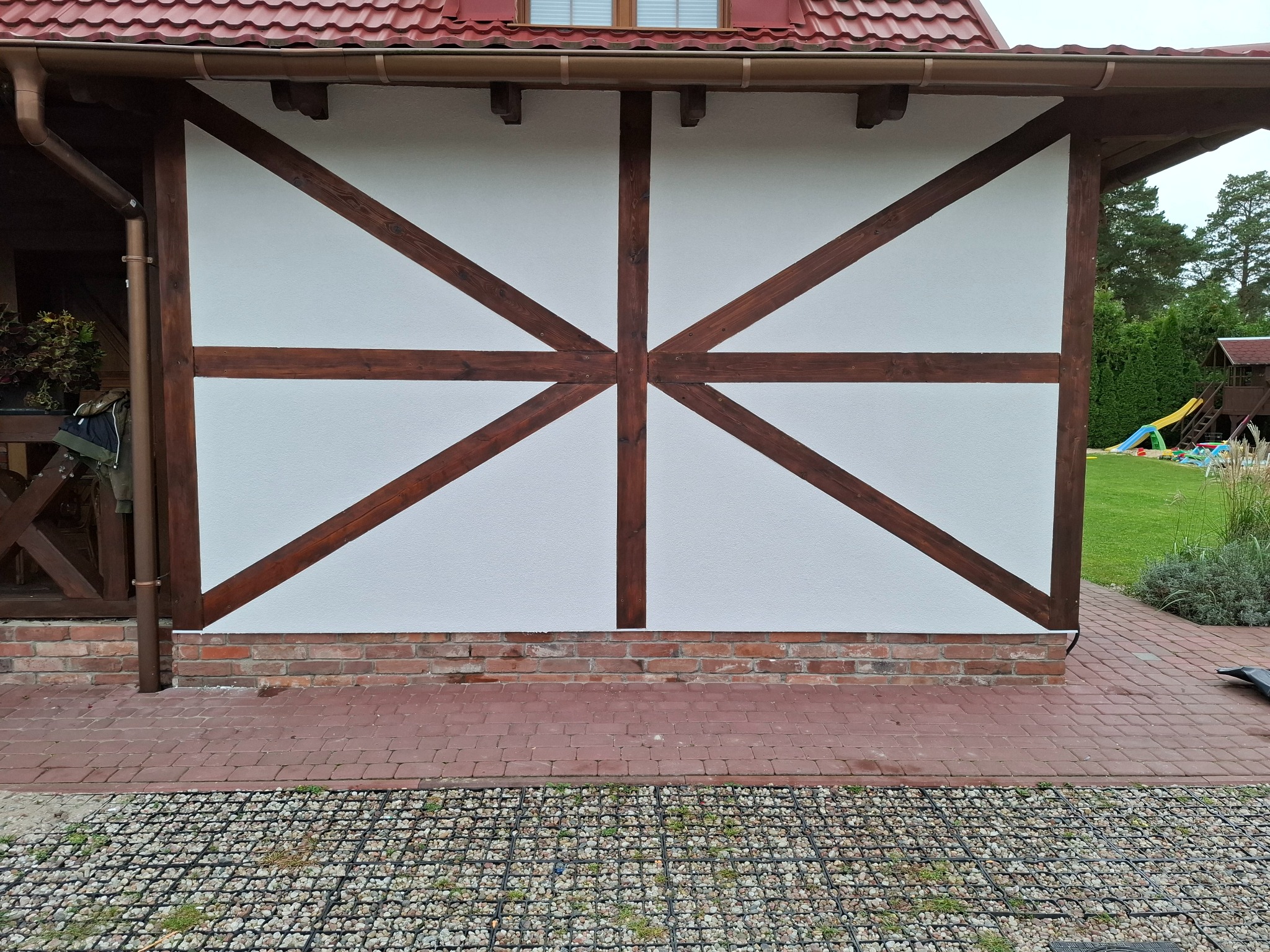 Biała ściana szachulcowa z brązowymi belkami, ceglana podmurówka i czerwona kostka brukowa. W tle trawnik i plac zabaw. Detal architektury.