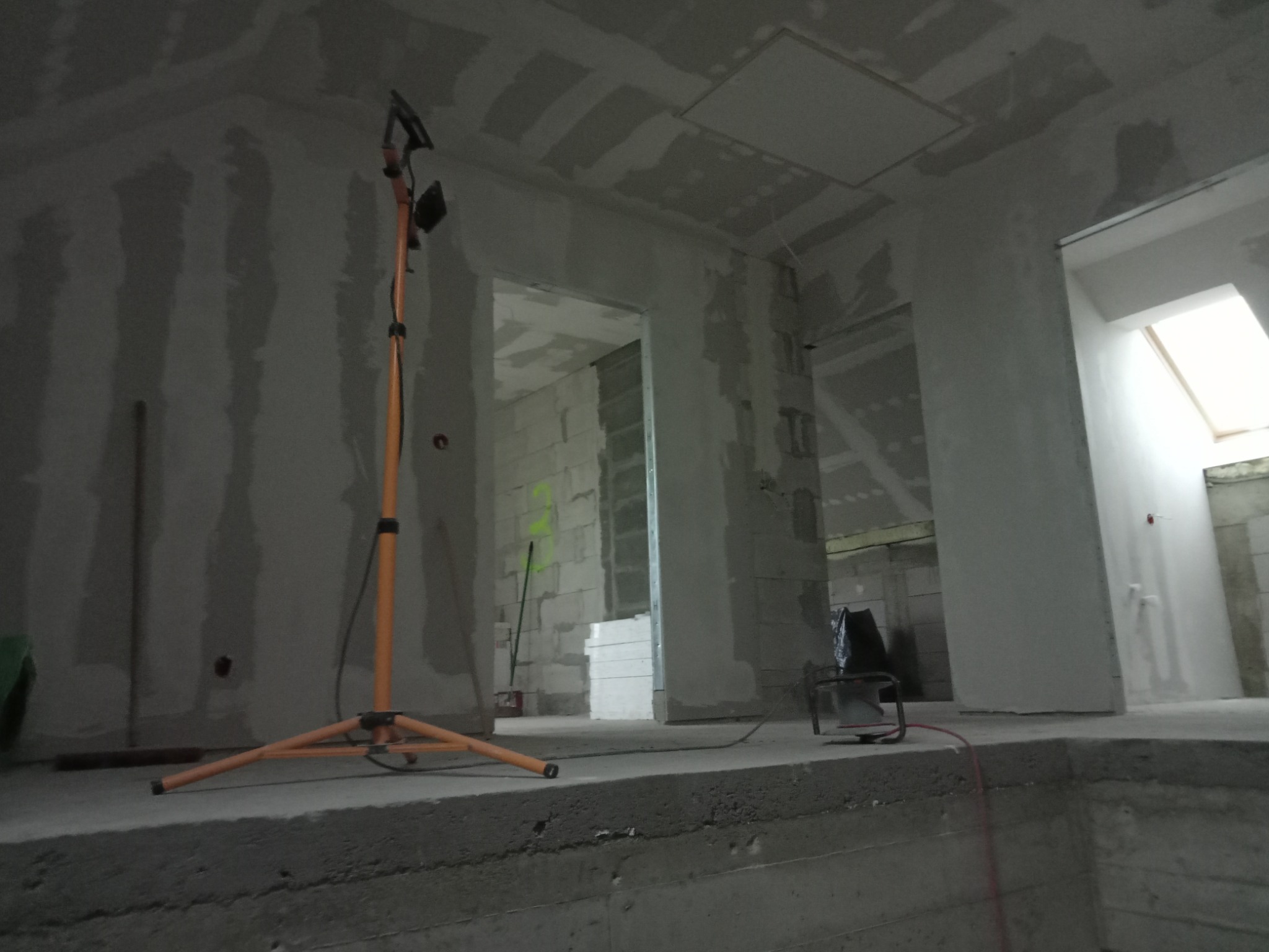 Wnętrze w trakcie prac wykończeniowych, widoczne ściany przygotowane pod malowanie z nałożoną warstwą gładzi, oświetlone przez lampę budowlaną na trójnogu. Widoczne surowe ściany z bloczków...