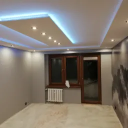 Pokój po remoncie z podwieszanym sufitem z oświetleniem LED, szarą ścianą z fototapetą górską i białym grzejnikiem pod drewnianym oknem.