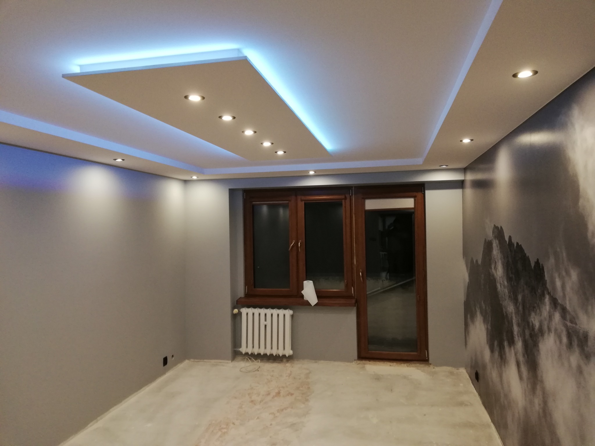 Pokój po remoncie z podwieszanym sufitem z oświetleniem LED, szarą ścianą z fototapetą górską i białym grzejnikiem pod drewnianym oknem.