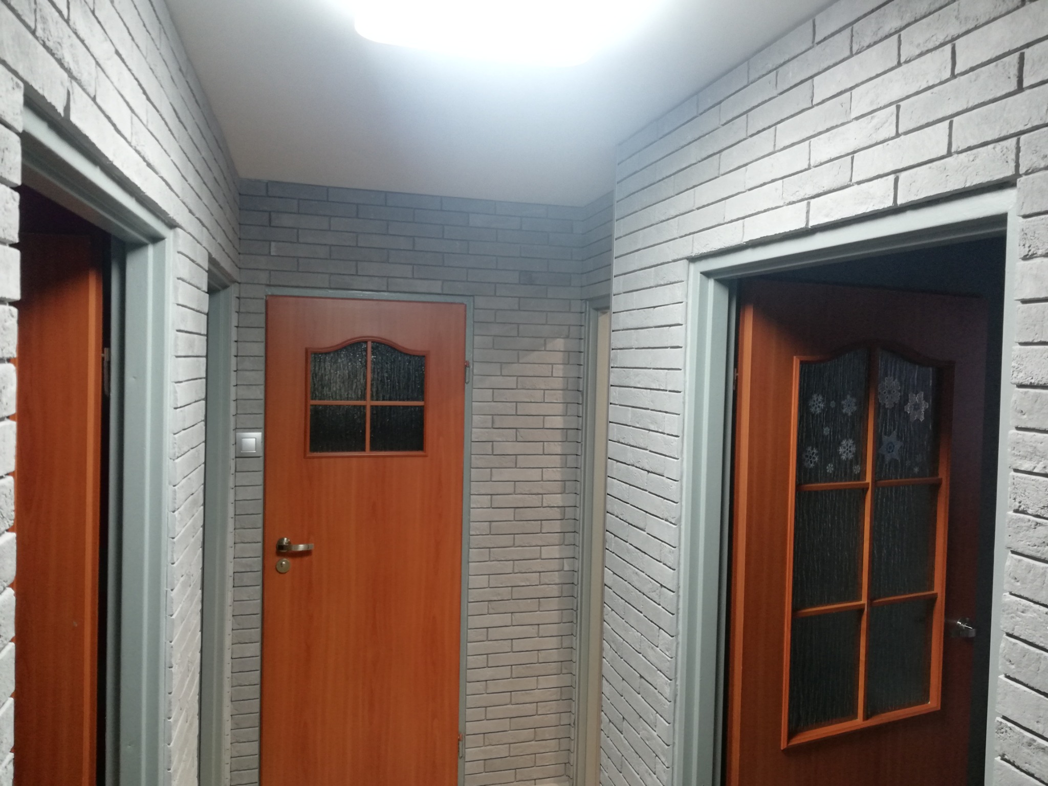 Korytarz wykończony dekoracyjną cegłą w odcieniach szarości, z dwiema drewnianymi drzwiami z przeszkleniami i szarą ościeżnicą, oświetlony górnym światłem.