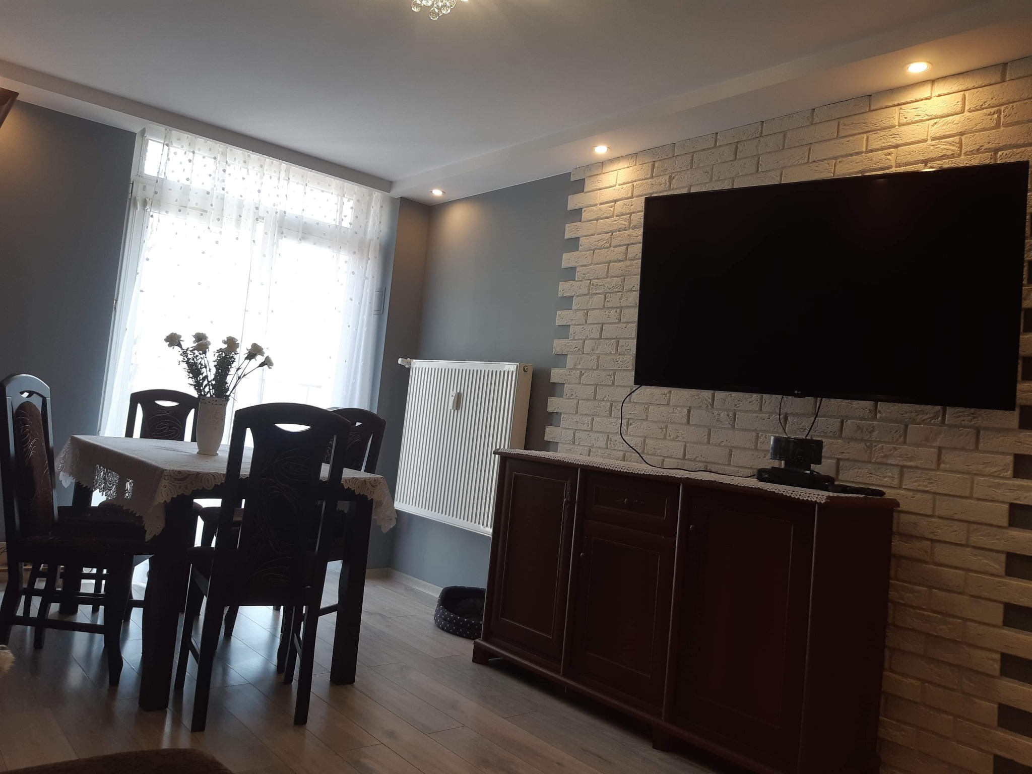 Jasne wnętrze salonu z ceglaną ścianą, ciemnymi meblami i stołem nakrytym koronkowym obrusem, oświetlone punktowymi lampami.