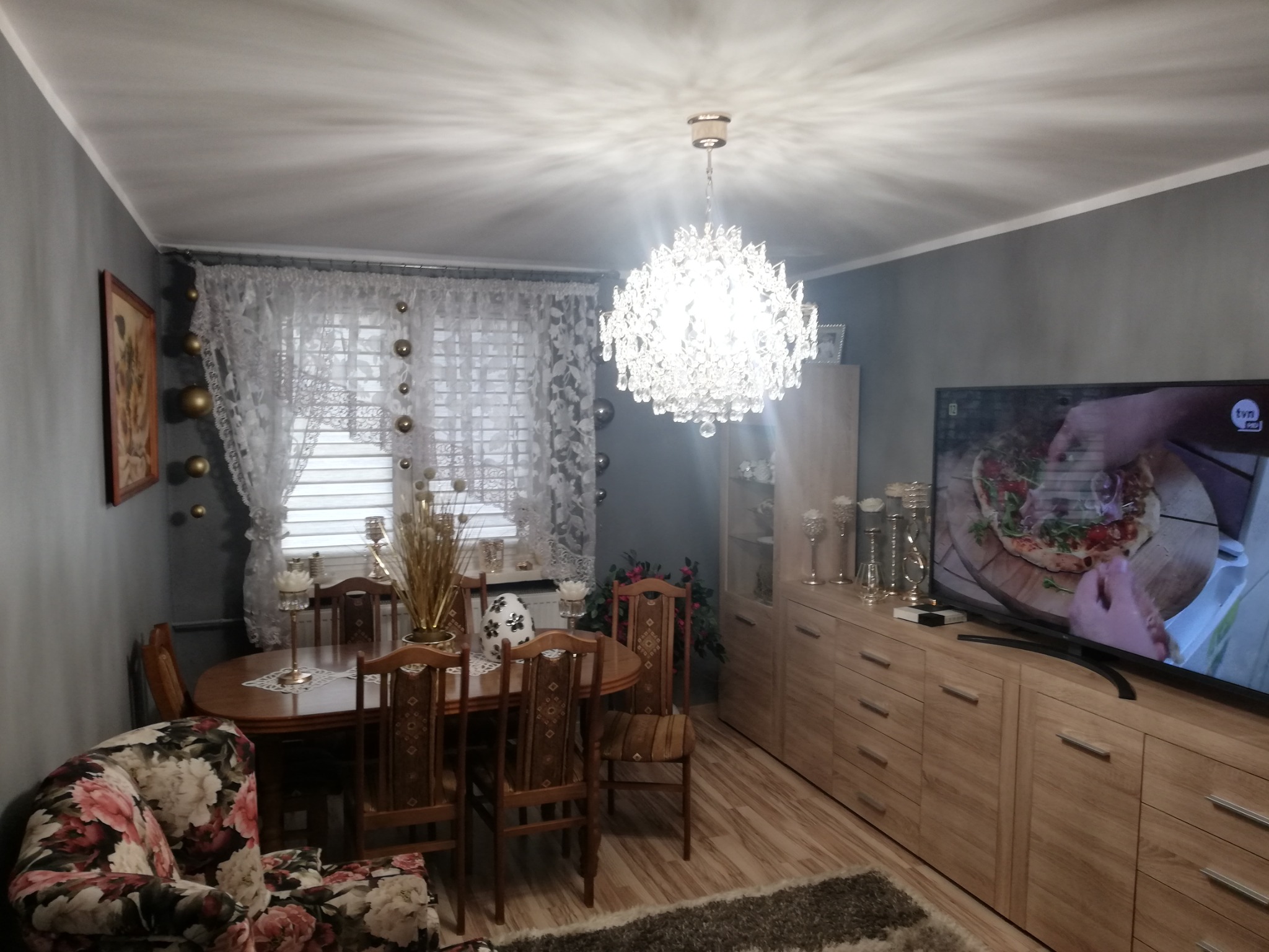 Jasne wnętrze jadalni z drewnianym stołem, krzesłami, kredensem i ozdobnym żyrandolem; na ścianach szara farba, na oknie białe firanki z dekoracjami.