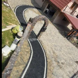 Ułożona kostka brukowa w kształcie łuku prowadzącego do domu, z elementami z kamienia i widocznymi workami z materiałami budowlanymi po bokach.