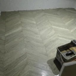 Ułożona pod kątem drewnopodobna podłoga z płytek, wiadro z narzędziami do fugowania w rogu, wzorzysta tapeta w górnej części ściany.