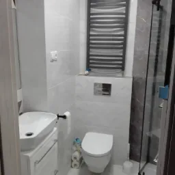 Nowoczesna łazienka z białymi płytkami o wzorze geometrycznym, wisząca umywalka z szafką, grzejnik drabinkowy i kabina prysznicowa z elementami imitującymi marmur.