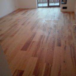 MS Bud - Jasne drewniane panele podłogowe ułożone w nowym pokoju z dużym oknem balkonowym i niedużym kominkiem.