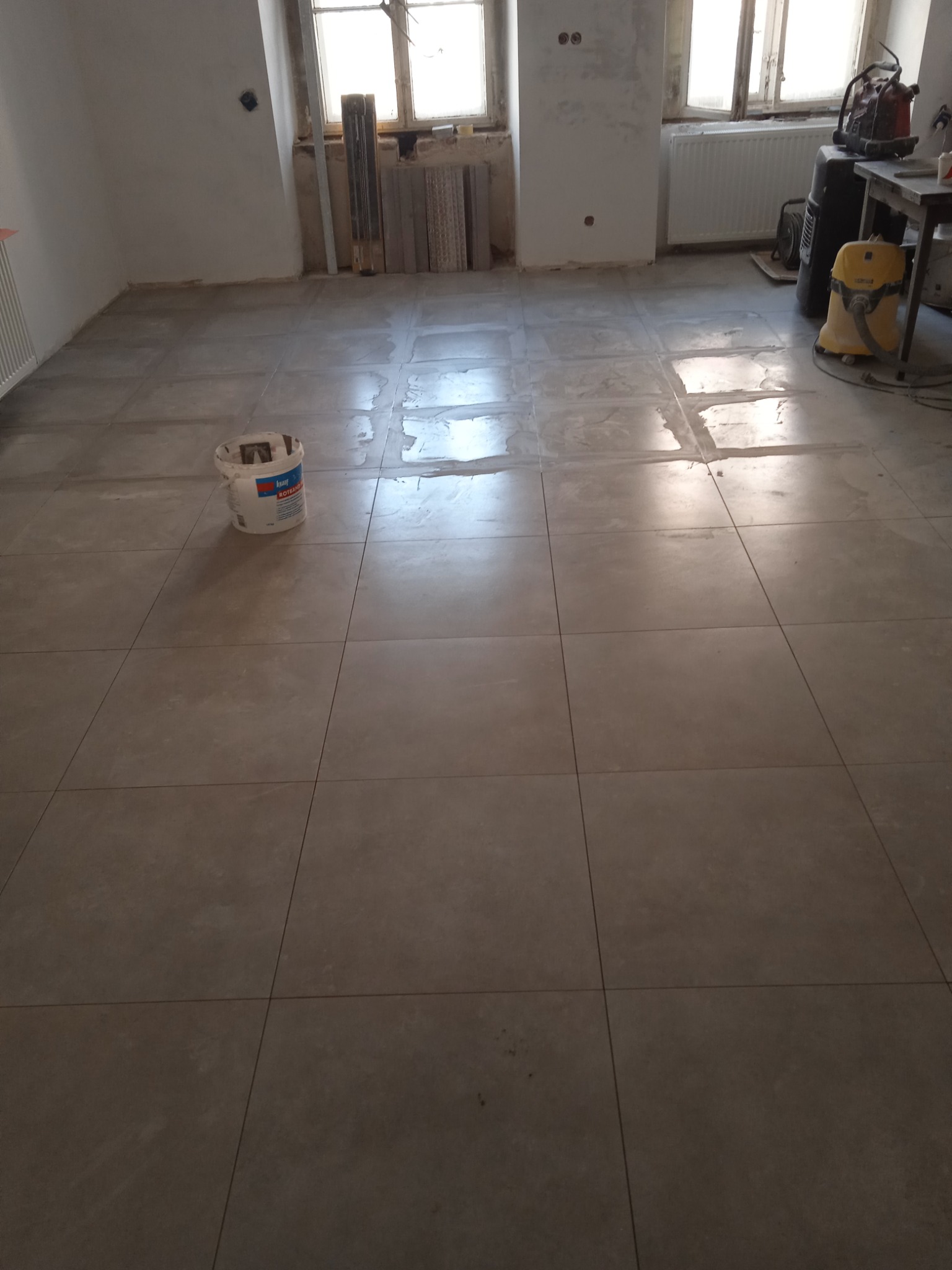 Ułożone szare płytki podłogowe w trakcie fugowania, widoczne narzędzia i wiadro z zaprawą, surowe ściany w tle.