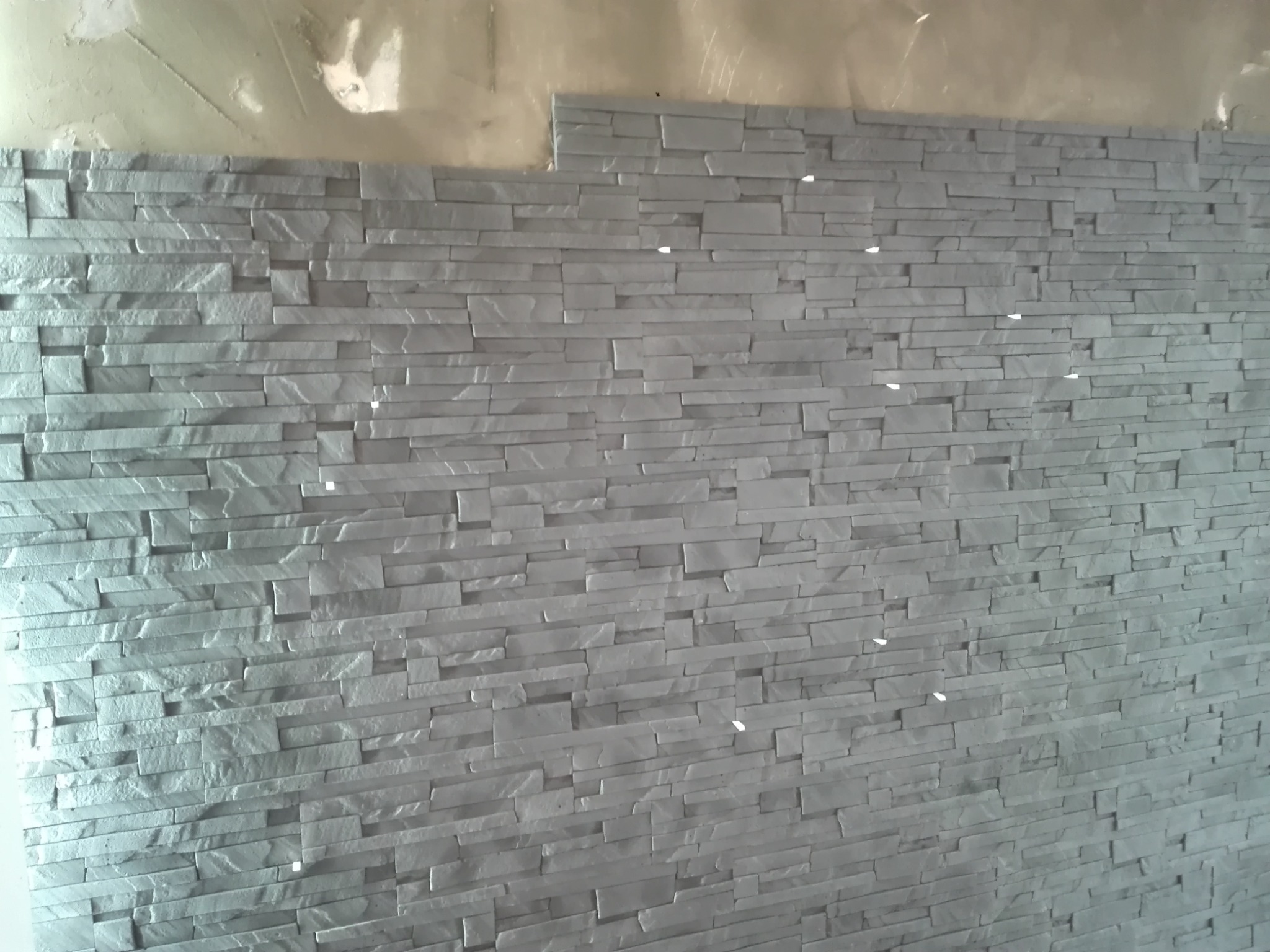 Ściana wyłożona szarymi, dekoracyjnymi płytkami imitującymi kamień, z widocznymi fragmentami surowej ściany powyżej.