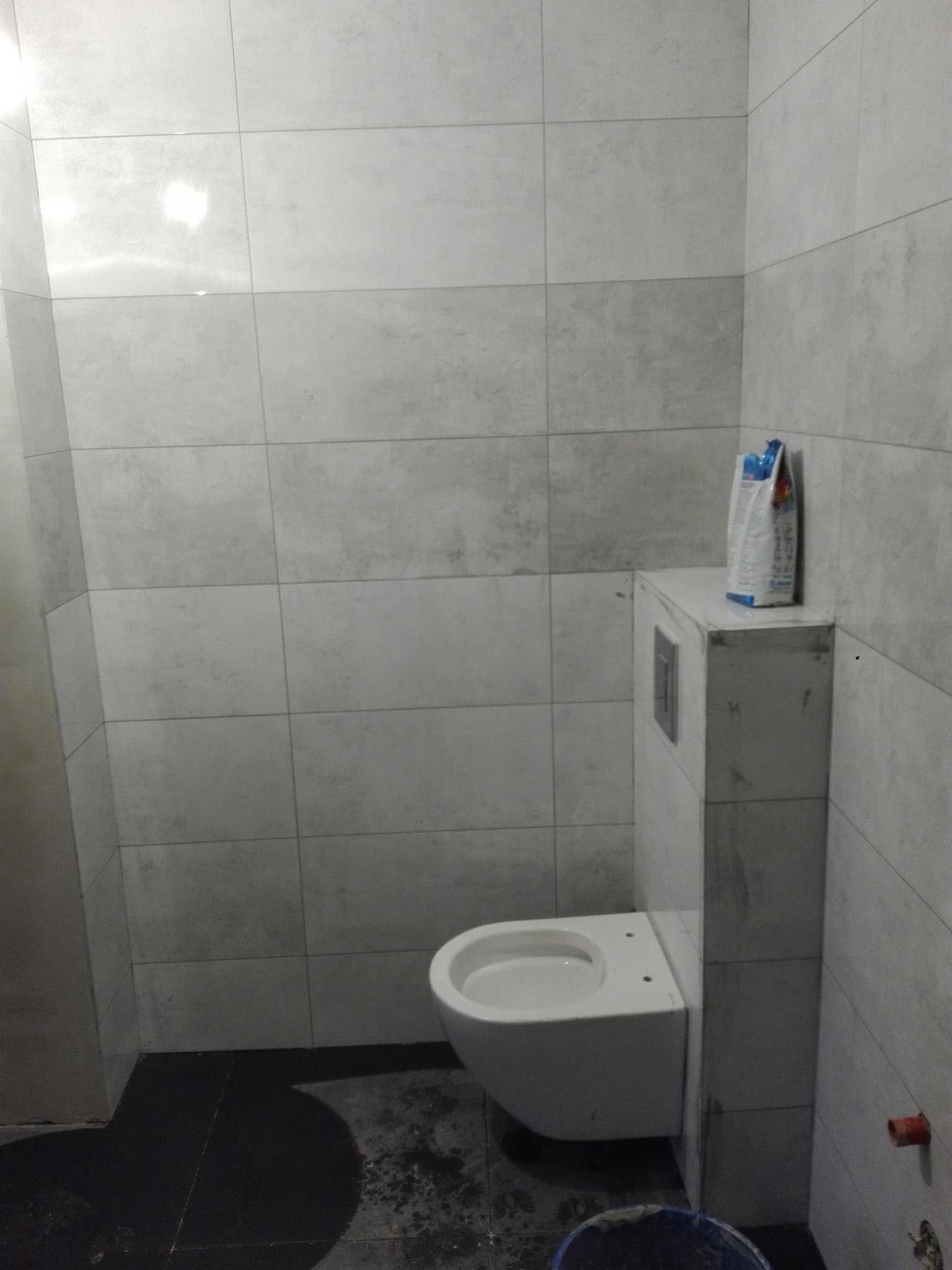 Wnętrze łazienki w trakcie remontu, widoczne szare płytki na ścianach, podwieszana toaleta, wiadro i torba z zaprawą na posadzce z ciemnych płytek.