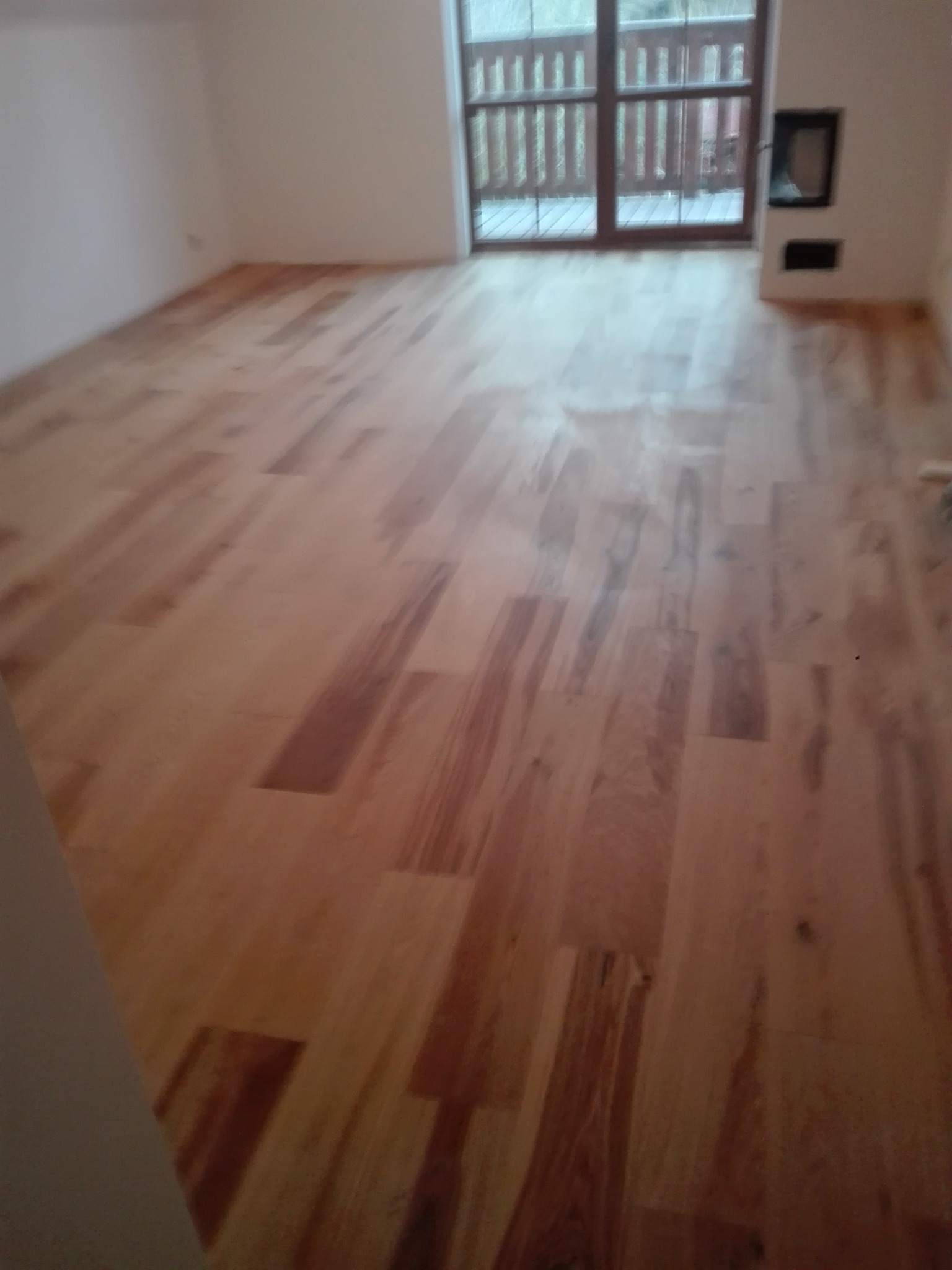 Jasne drewniane panele podłogowe ułożone w nowym pokoju z dużym oknem balkonowym i niedużym kominkiem.