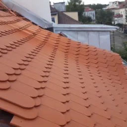 Nowe, pomarańczowe dachówki karpiówki ułożone na dachu budynku mieszkalnego, widok z góry na fragment dachu z kominem i budynkami w tle.