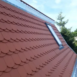 Nowy dach pokryty czerwoną dachówką karpiówką z zamontowanym oknem dachowym i obróbką blacharską, widok z dołu na tle błękitnego nieba i zieleni drzew.