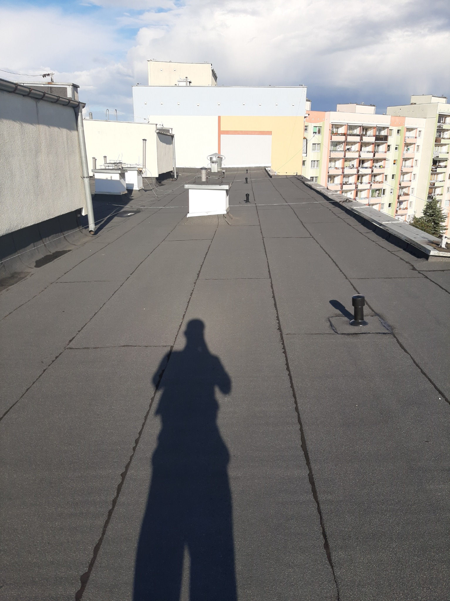 Bitumiczna płaska powierzchnia dachu z widocznym cieniem fotografa, wentylacją i budynkami w tle.