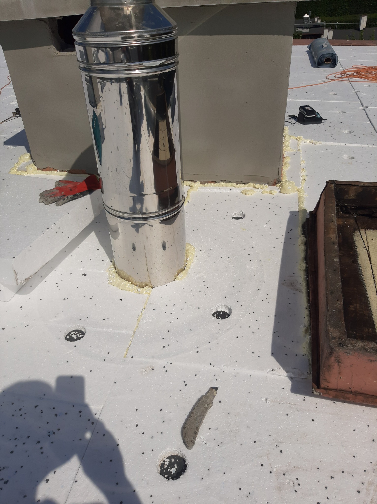 Montaż komina systemowego na izolacji dachu płaskiego, widoczne uszczelnienie pianką montażową, otwory wentylacyjne w izolacji, rękawice robocze i cień fotografa.