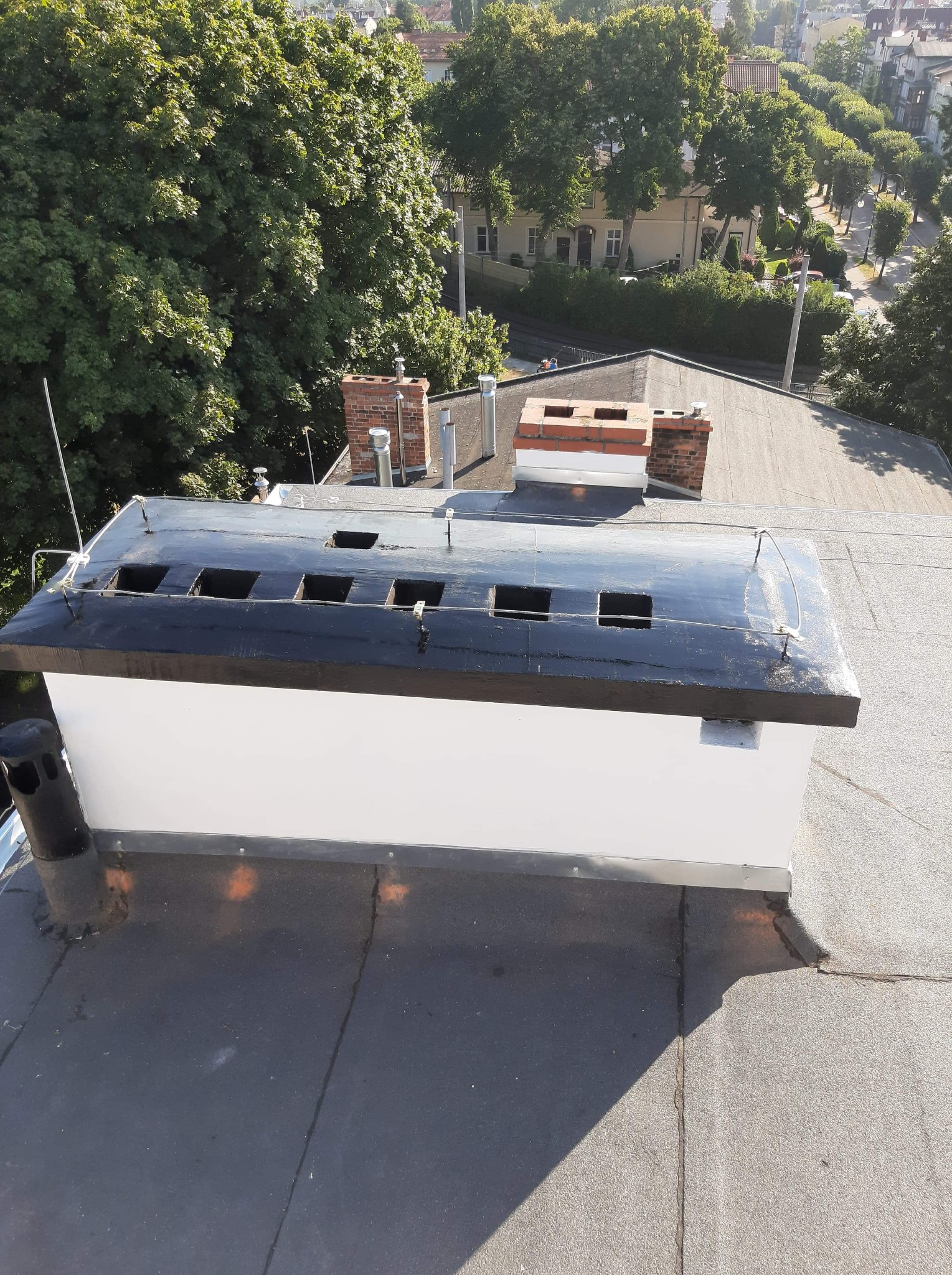 Widok z góry na fragment dachu pokrytego papą z kominem wentylacyjnym z czarną, błyszczącą obudową i otworami wentylacyjnymi, na tle zieleni drzew i zabudowań miejskich.