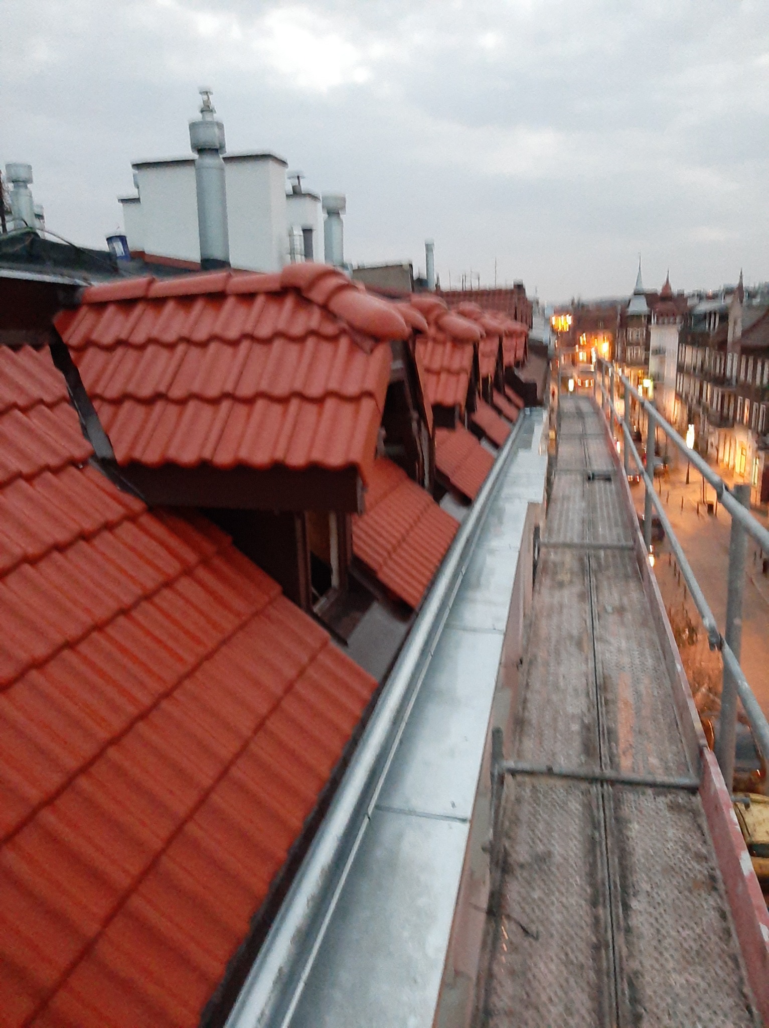 Widok z rusztowania na dach z czerwonej dachówki i nową, błyszczącą rynnę; w tle zabudowa miejska oświetlona wieczornym światłem.