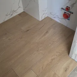 Ułożona podłoga z płytek imitujących drewno w łazience, białe marmurowe płytki na ścianach, widoczne rury instalacji wodnej w ścianie.