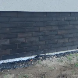 Fragment ściany budynku z ciemnej cegły klinkierowej, widoczna warstwa izolacji przy gruncie i fragment elewacji w jasnym kolorze.