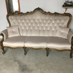 Elegancka, beżowa sofa w stylu Ludwika, z pikowanym oparciem zdobionym kryształkami i drewnianą ramą w kolorze złota, stoi na betonowej podłodze. Na oparciu widoczne dwie poduszki. W tle ściana...