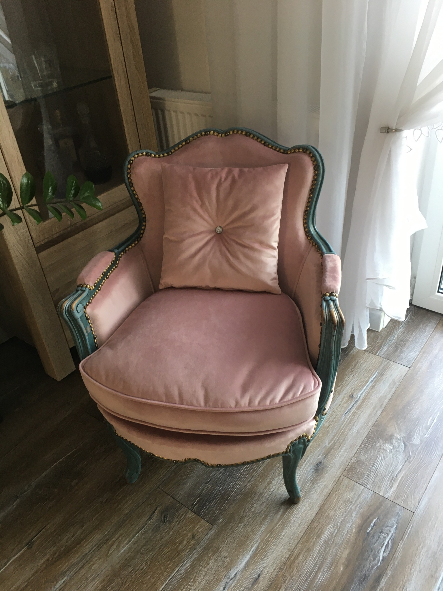 Odrestaurowany fotel w stylu vintage z różowym welurem i turkusowymi zdobieniami, z poduszką z guzikiem, na drewnianej podłodze.