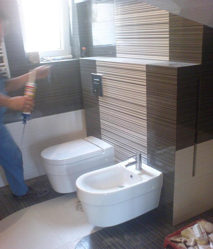 Montaż armatury w łazience z brązowo-białymi płytkami: biała toaleta i bidet, osoba w niebieskim kombinezonie uszczelnia silikonem szczelinę przy ścianie.