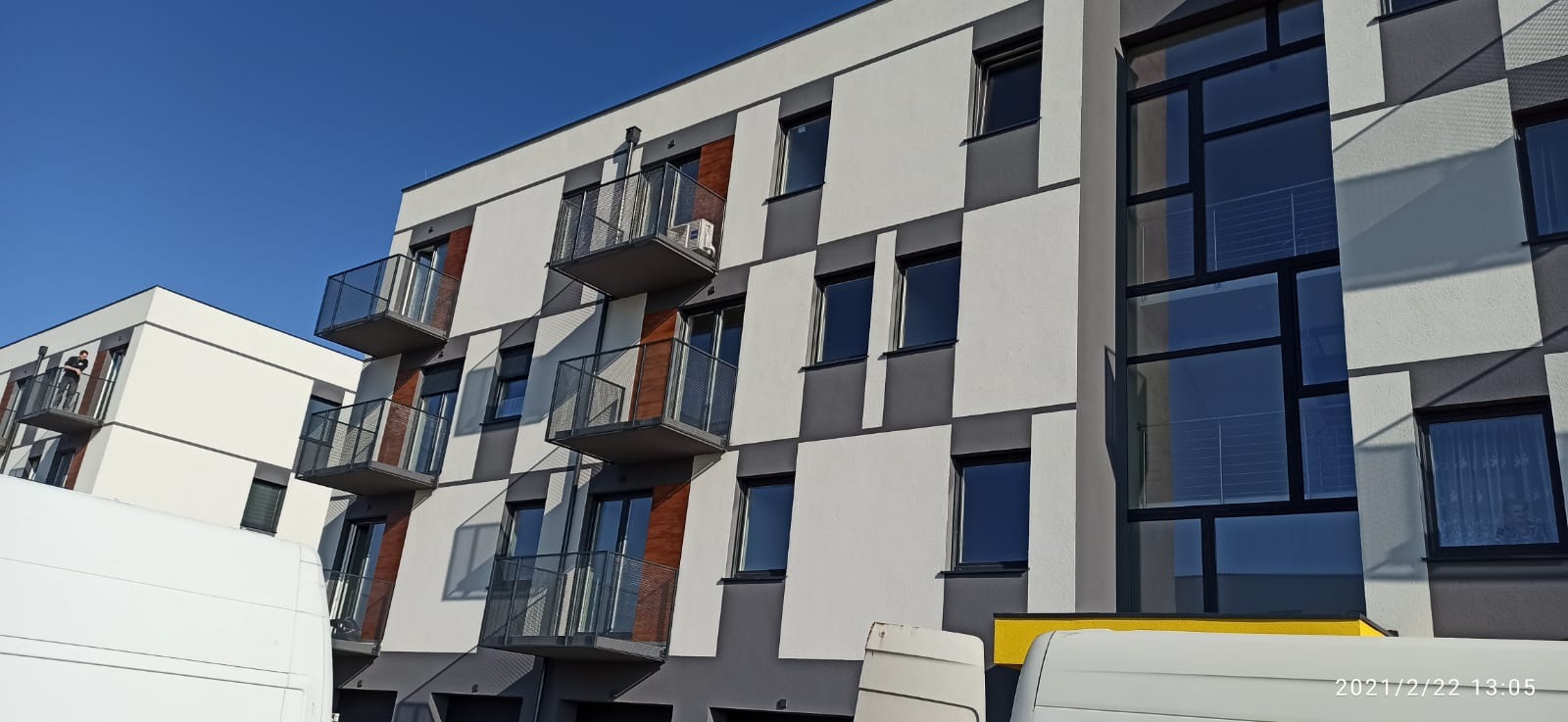 Nowoczesny budynek wielorodzinny z balkonami, z widoczną jednostką zewnętrzną klimatyzacji na jednym z nich, fasada w geometryczne wzory bieli i szarości, fragment białego samochodu dostawczego...