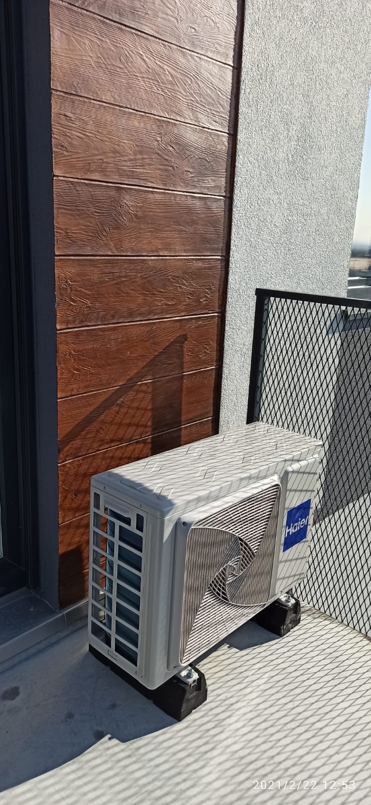 Zewnętrzna jednostka klimatyzacji Haier zamontowana na balkonie, widoczna na gumowych podkładkach antywibracyjnych, z brązową ścianą imitującą drewno i szarą elewacją w tle.