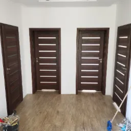 Wnętrze korytarza z trzema zamontowanymi, nowoczesnymi drzwiami w odcieniach brązu, z widocznymi śladami po montażu: wiadro z narzędziami i śmieci na podłodze.