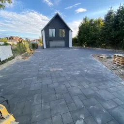 Ułożona, ciemnoszara kostka brukowa prowadząca do nowoczesnego domu z ciemną elewacją, widoczna ubijarka w rogu kadru, błękitne niebo z chmurami w tle.