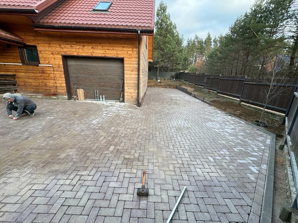 Pracownik w szarej bluzie klęczy na nowo ułożonej, wzorzystej nawierzchni z kostki brukowej, wyrównując fugi; w tle drewniany budynek z brązową bramą garażową i las.