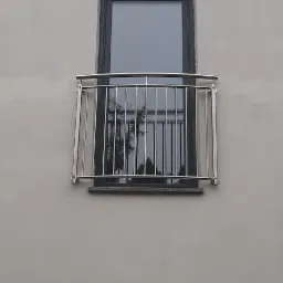 Nierdzewna balustrada balkonowa zamontowana przy oknie na elewacji budynku o szarej, surowej fakturze. W szybie okna widoczne odbicie drzew.