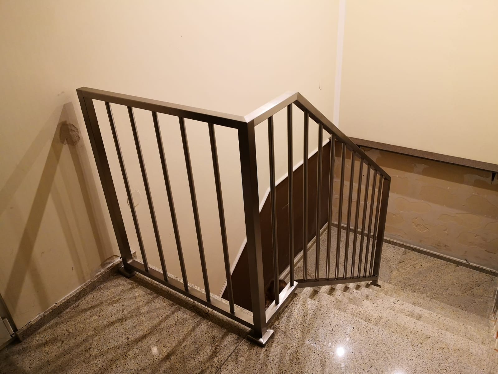 Schody z zamontowaną, metalową balustradą w kolorze ciemnego brązu, widok z góry na dół schodów, ściany w kolorze kremowym, podłoga wyłożona płytkami imitującymi lastryko.