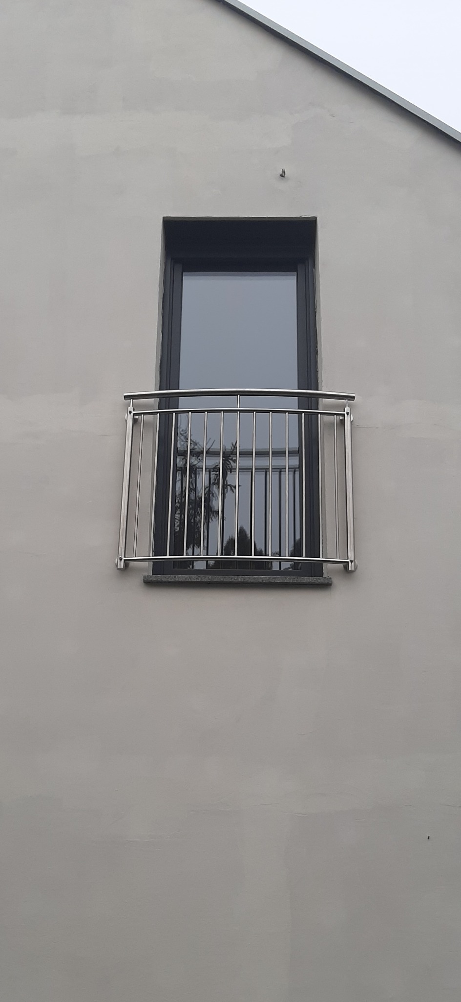 Nierdzewna balustrada balkonowa zamontowana przy oknie na elewacji budynku o szarej, surowej fakturze. W szybie okna widoczne odbicie drzew.