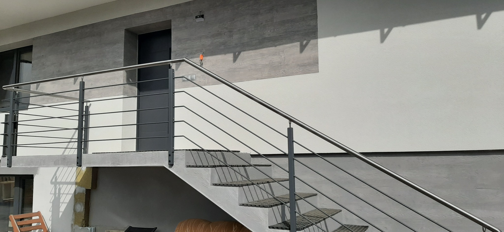 Zewnętrzne schody z balustradą ze stali nierdzewnej z poziomymi prętami, widoczne elementy montażowe, szare stopnie i ściany budynku w odcieniach szarości i bieli.