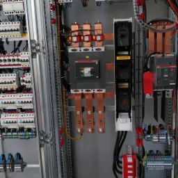 Rozdzielnica elektryczna z widocznymi bezpiecznikami, wyłącznikami, szynami miedzianymi i okablowaniem, zamontowana w metalowej obudowie z szarymi ściankami.