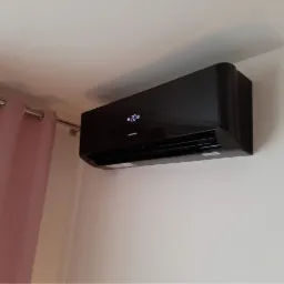 Czarny klimatyzator ścienny włączony, wyświetla temperaturę 20 stopni, widoczny fragment różowej zasłony.