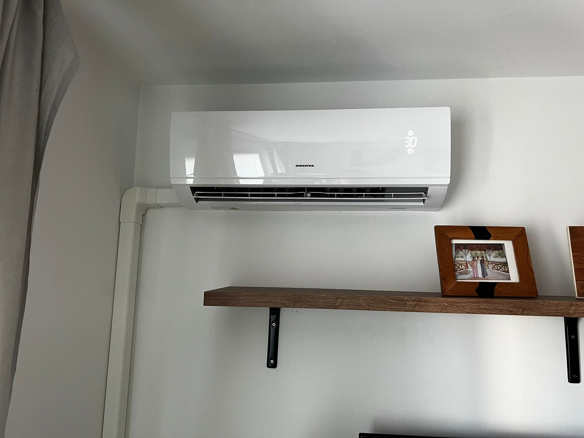 Biała klimatyzacja Innova z wyświetlaczem temperatury 30 stopni Celsjusza, zamontowana na ścianie nad drewnianą półką z ramkami na zdjęcia.