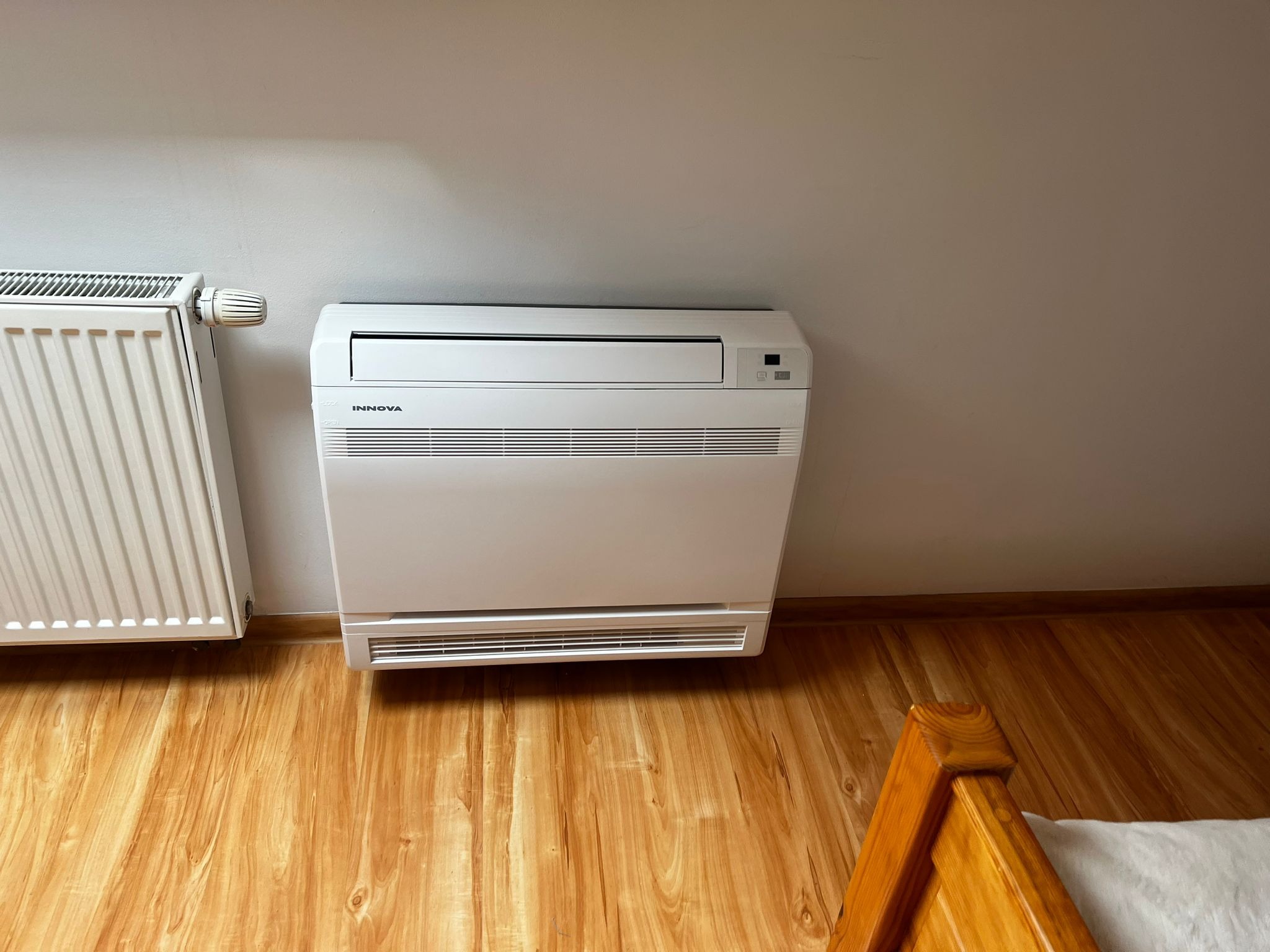 Biały klimatyzator podłogowy marki Innova zamontowany na ścianie, obok grzejnika, na tle drewnianej podłogi.