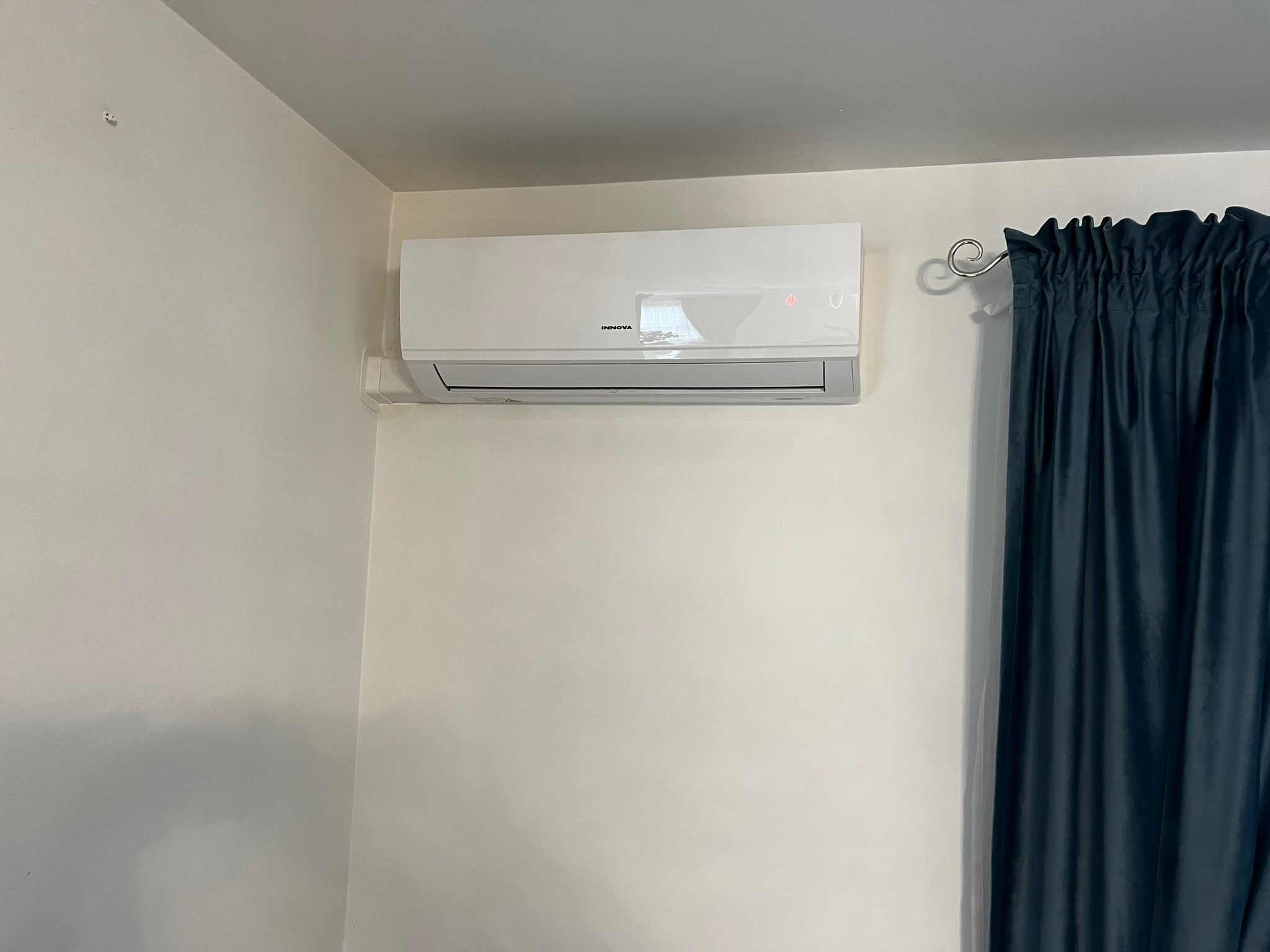 Biały klimatyzator marki Innova zamontowany na jasnej ścianie w rogu pokoju, obok ciemnozielonej zasłony z ozdobnym metalowym karniszem.