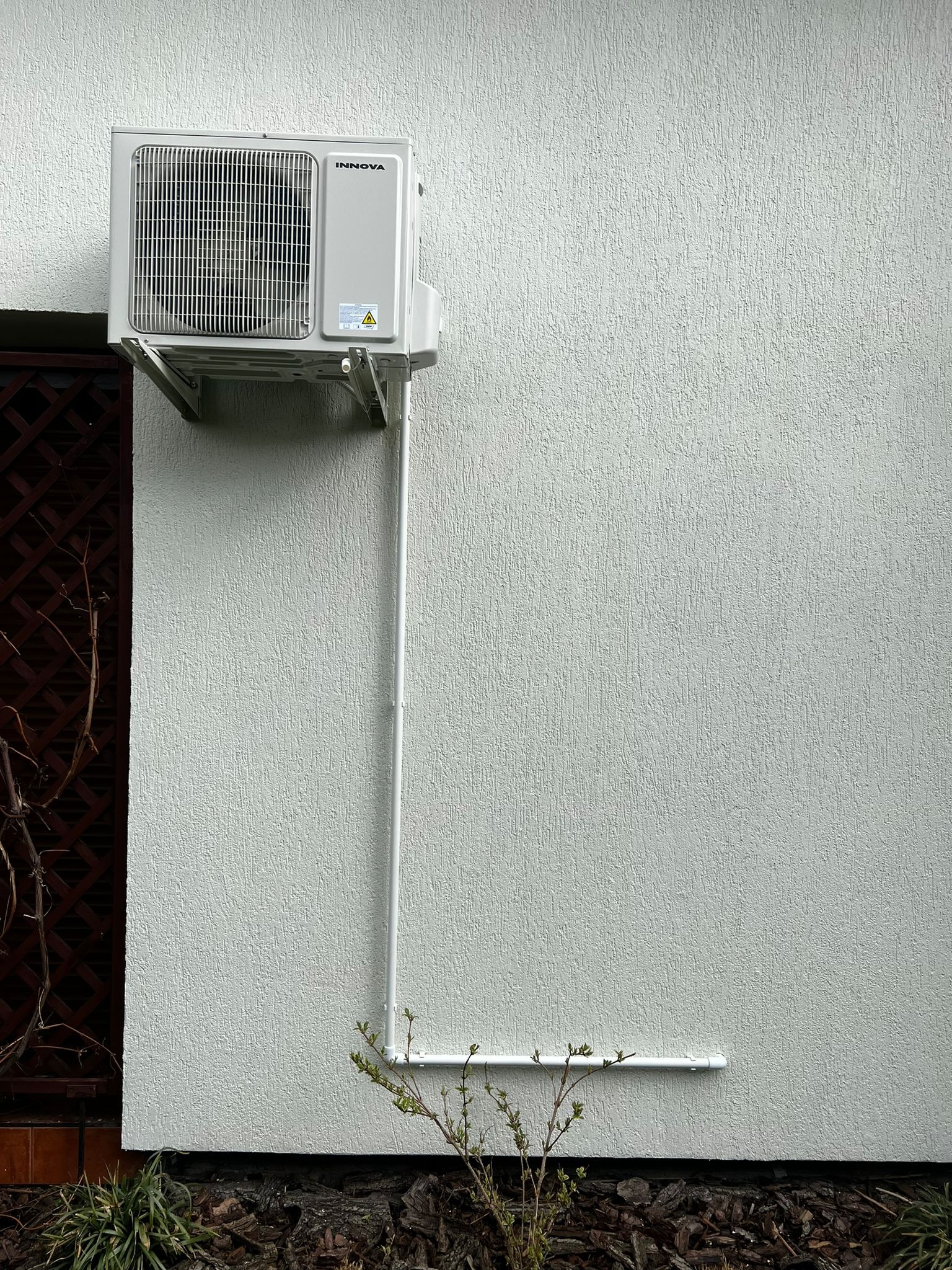 Zewnętrzna jednostka klimatyzacji marki Innova zamontowana na teksturowanej, jasnej ścianie budynku, z widoczną instalacją odprowadzającą skropliny, umieszczoną przy brązowej kratownicy i małej...