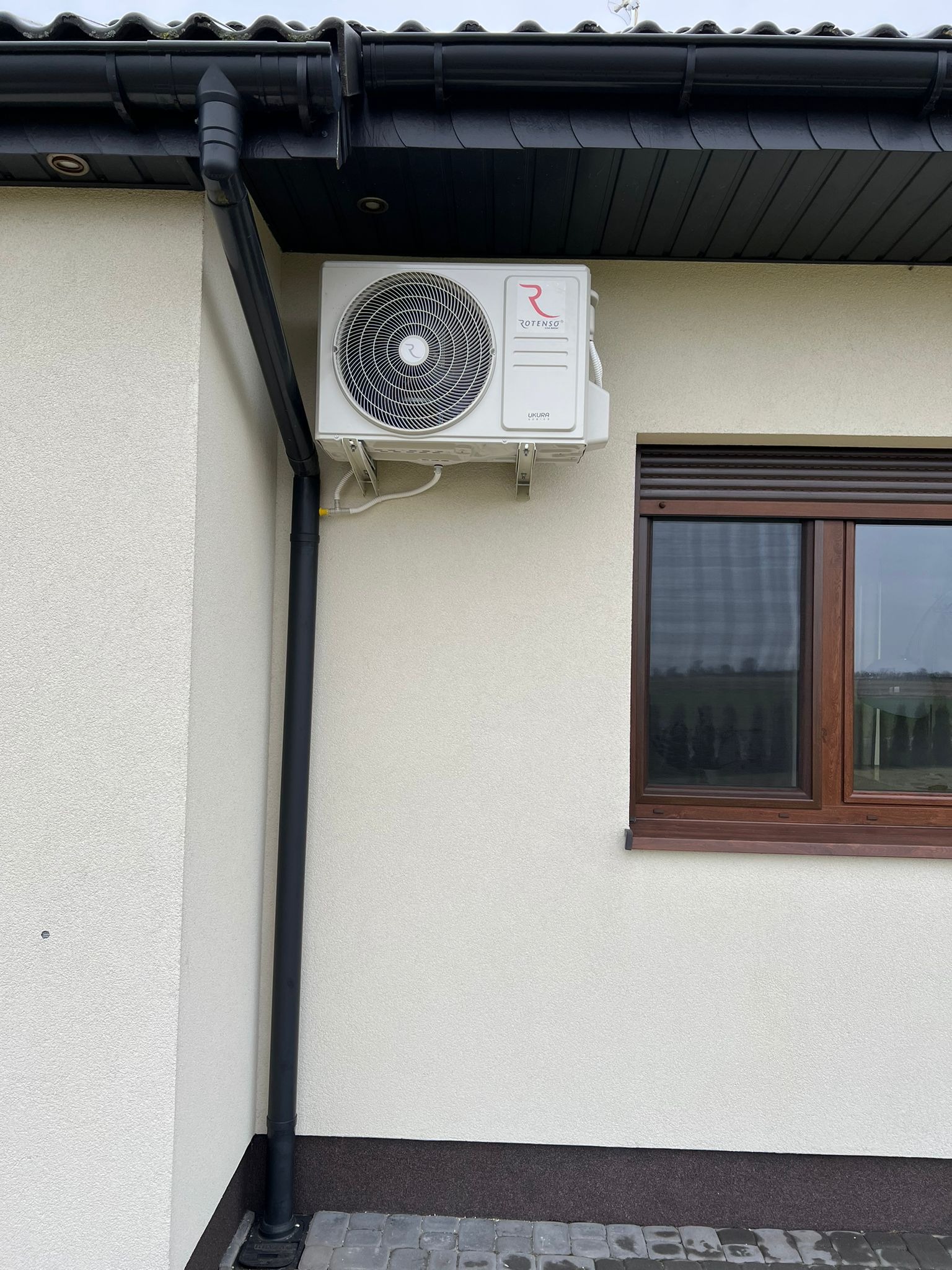 Biała jednostka zewnętrzna klimatyzacji Rotenso Ukura zamontowana na kremowej elewacji domu obok brązowego okna z roletami i czarnej rynny spustowej.
