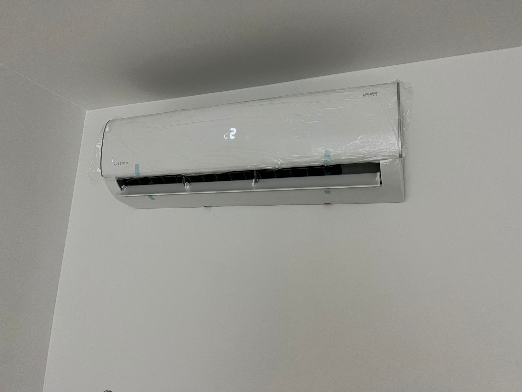 Nowy, biały klimatyzator marki Rotenso, jeszcze w folii ochronnej, zamontowany na ścianie. Na wyświetlaczu widoczna temperatura 22 stopnie.